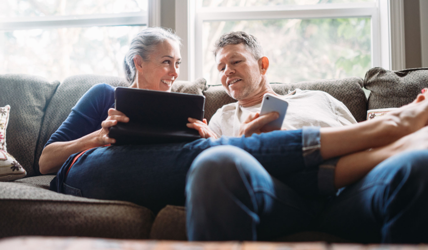 Un couple détendu à la maison, consultant une tablette et un smartphone, illustrant la simplicité et l'accessibilité des solutions de facturation de chauffage à distance proposées par Groupe E pour les clients privés