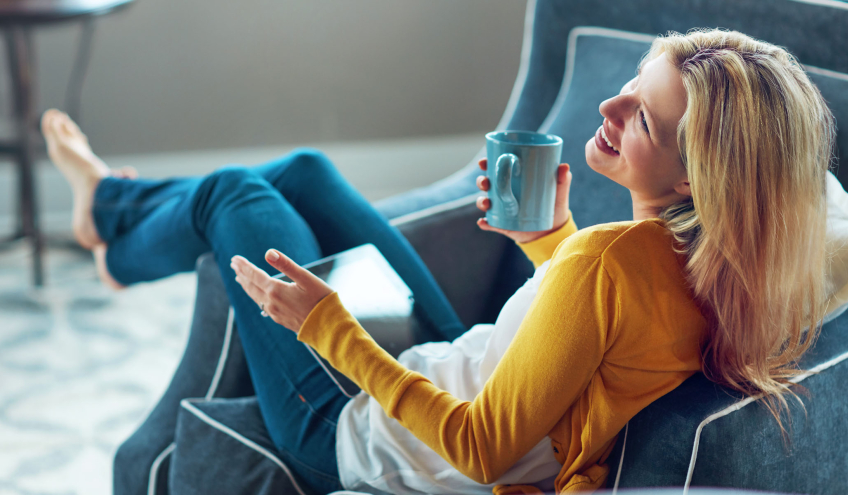 Une femme souriante, détendue dans un fauteuil, tenant une tasse, symbolisant le confort et la sérénité offerts par les solutions énergétiques de Groupe E