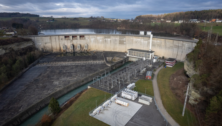 Luftaufnahme eines Wasserkraftwerks und seiner umliegenden elektrischen Anlagen, die die imposante Struktur und das Wasserbecken im Hintergrund zeigen. Dieser Standort veranschaulicht die Infrastruktur zur Erzeugung erneuerbarer Energie, ein Bereich, in dem Groupe E tätig ist, um nachhaltigen und umweltfreundlichen Strom bereitzustellen.