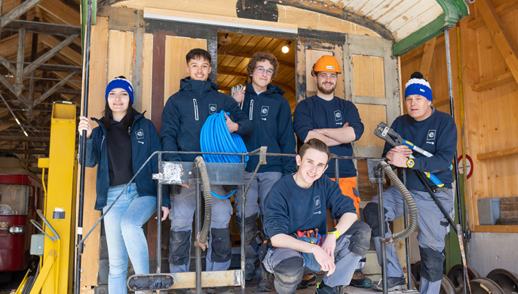 Un groupe de jeunes apprentis du Groupe E posant ensemble sur le marchepied d'un ancien tramway en rénovation. Chacun porte une tenue de travail avec le logo du Groupe E, et certains tiennent des outils ou du matériel électrique. Ils semblent souriants et motivés, reflétant l'engagement du Groupe E dans la formation professionnelle et la préservation du patrimoine. L'atelier en bois, en arrière-plan, donne un aspect chaleureux à l'environnement de travail.