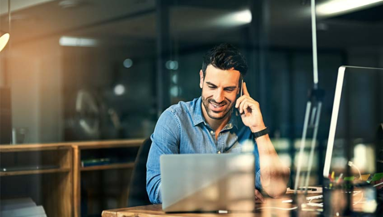 Un collaborateur de Groupe E, assis à son bureau, utilise son ordinateur portable tout en parlant au téléphone, souriant et concentré dans un environnement de bureau moderne.