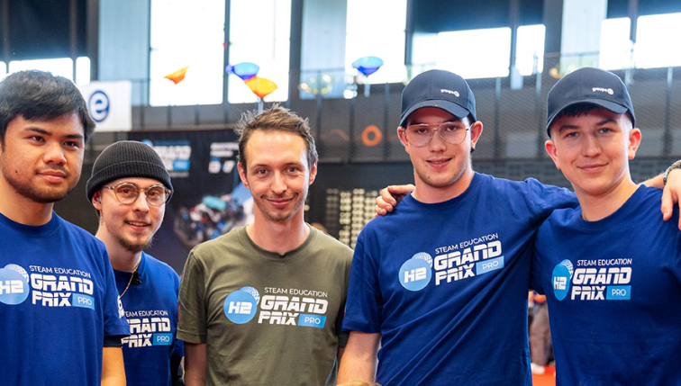 Eine Gruppe junger Teilnehmer am H2 Steam Education Grand Prix, organisiert von Groupe E, posiert gemeinsam in einer dynamischen und freundlichen Umgebung. Die Teilnehmer, gekleidet in T-Shirts und Kappen mit den Logos der Veranstaltung und von Groupe E, zeigen ihre Begeisterung für die Initiative. Im Hintergrund sind weitere Teilnehmer und bunte schwebende Elemente zu sehen, was die Energie und Innovation veranschaulicht, die Groupe E im Bereich Bildung und Technologie fördert.