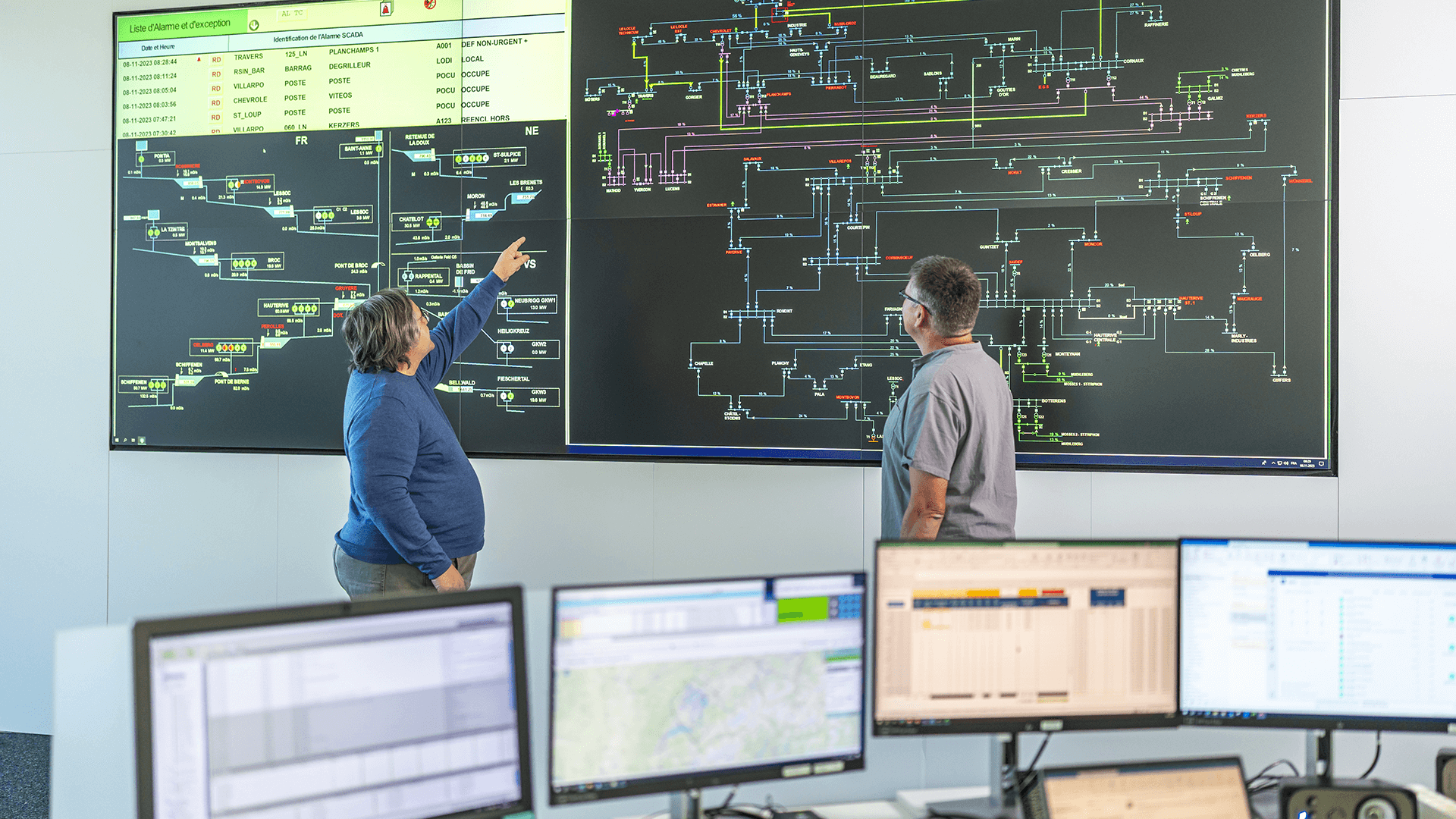 Texte alternatif : Deux collaborateurs de Groupe E analysent et supervisent un tableau de contrôle complexe affiché sur un grand écran, montrant le réseau de distribution énergétique. Ils travaillent dans un centre de dispatching moderne, entourés de plusieurs écrans d'ordinateurs, illustrant la gestion avancée de l'automation énergétique.